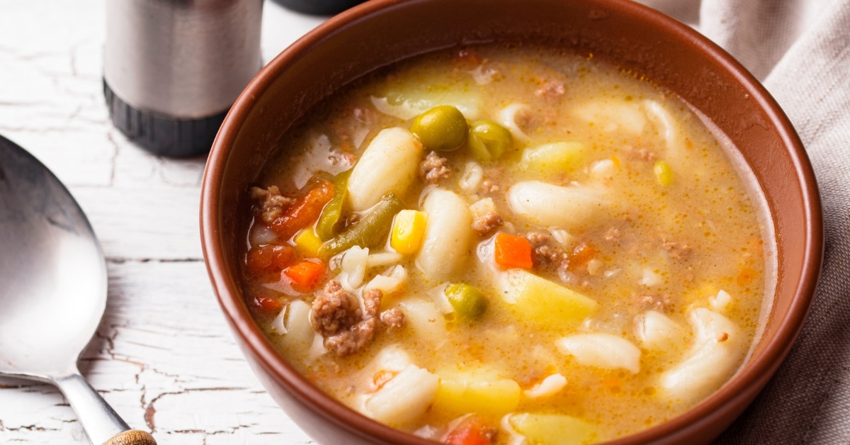 Busy day soup on a brown bowl made with elbow macaroni, green peas, carrots and broth.