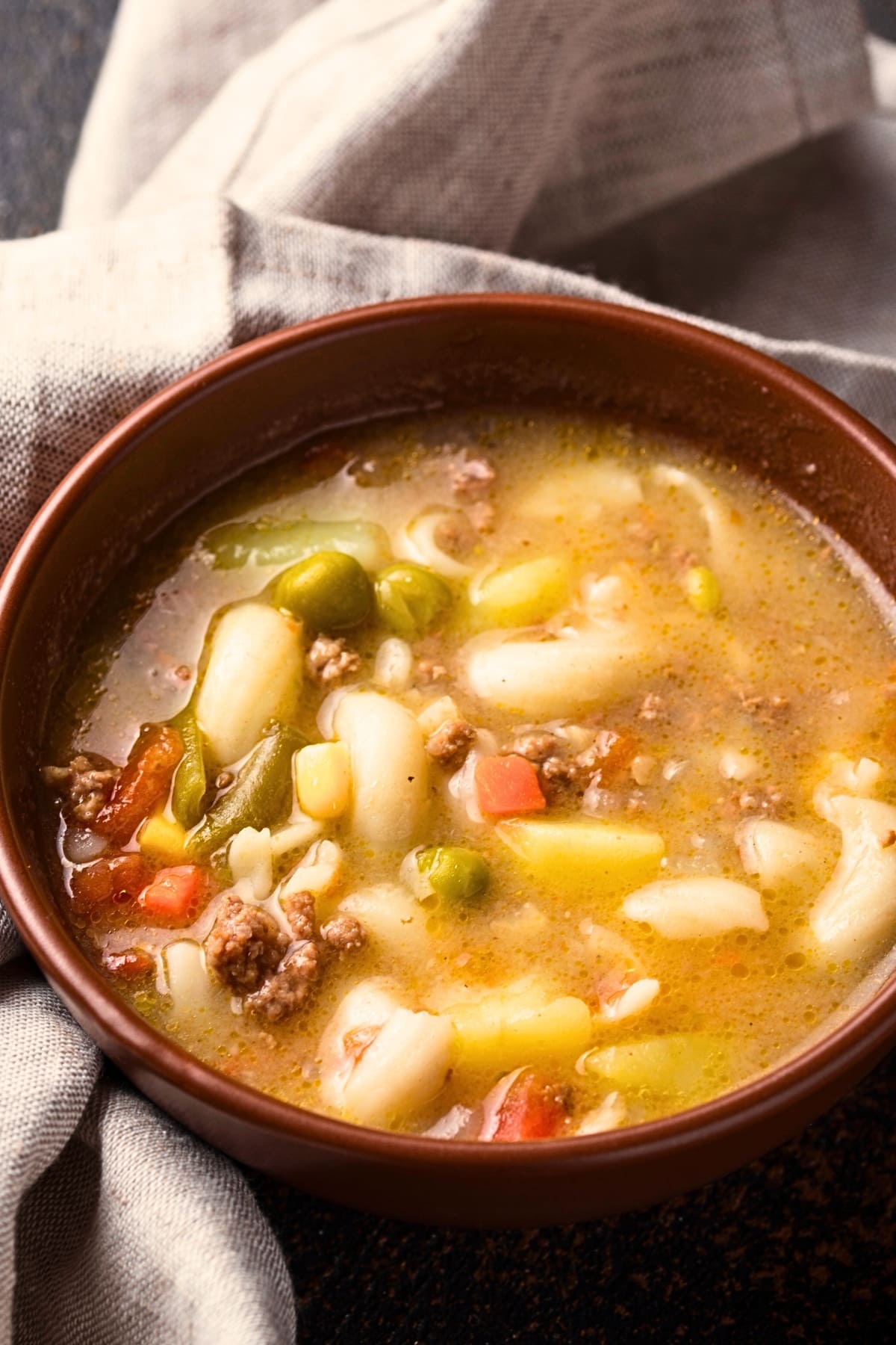 Busy day soup with elbow macaroni, ground meat, carrots, green peas and broth served on a brown bowl. 