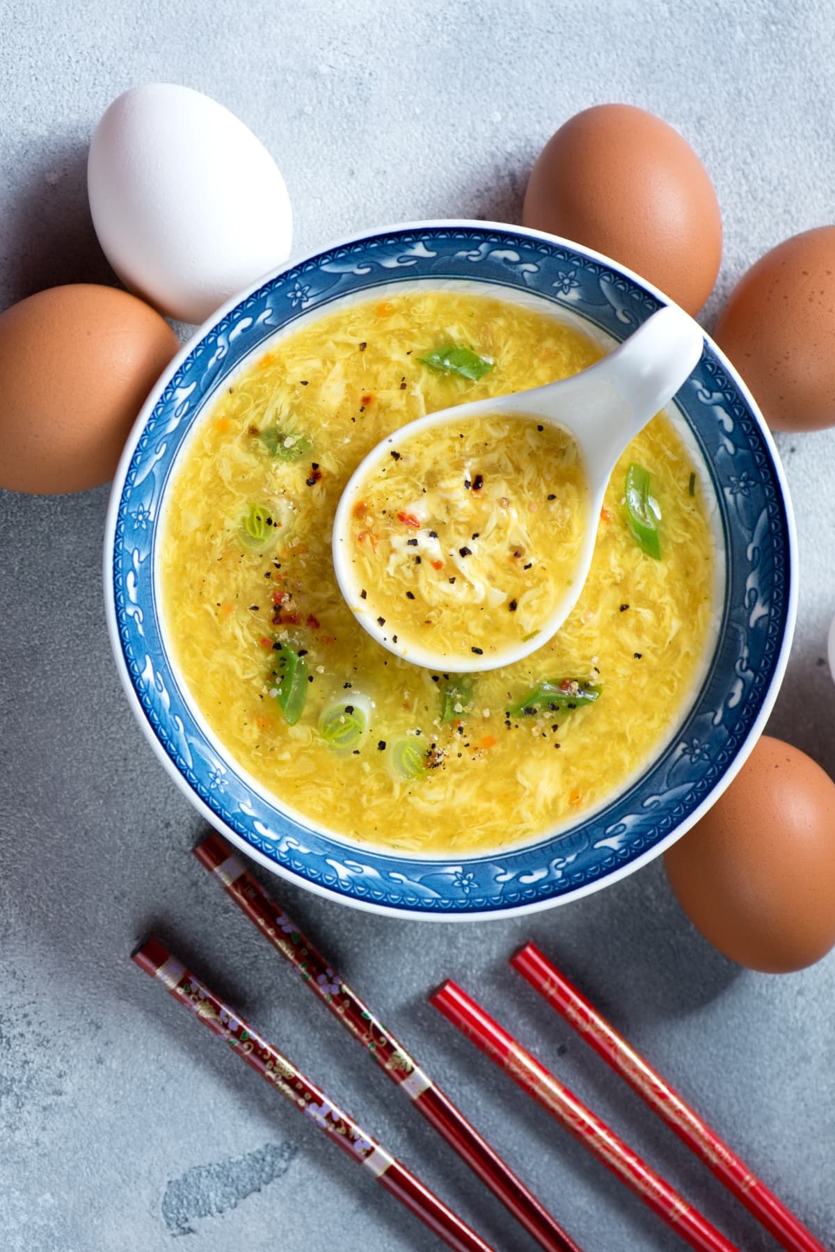 A Chinese bowl with egg drop soup garnished with chopped green onions and seasonings with chopsticks and raw eggs around it.