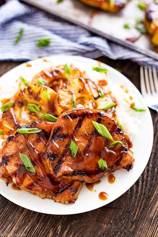 Grilled pork chop served with pineapple rings and sauce garnished with chopped chives served on a white plate. 