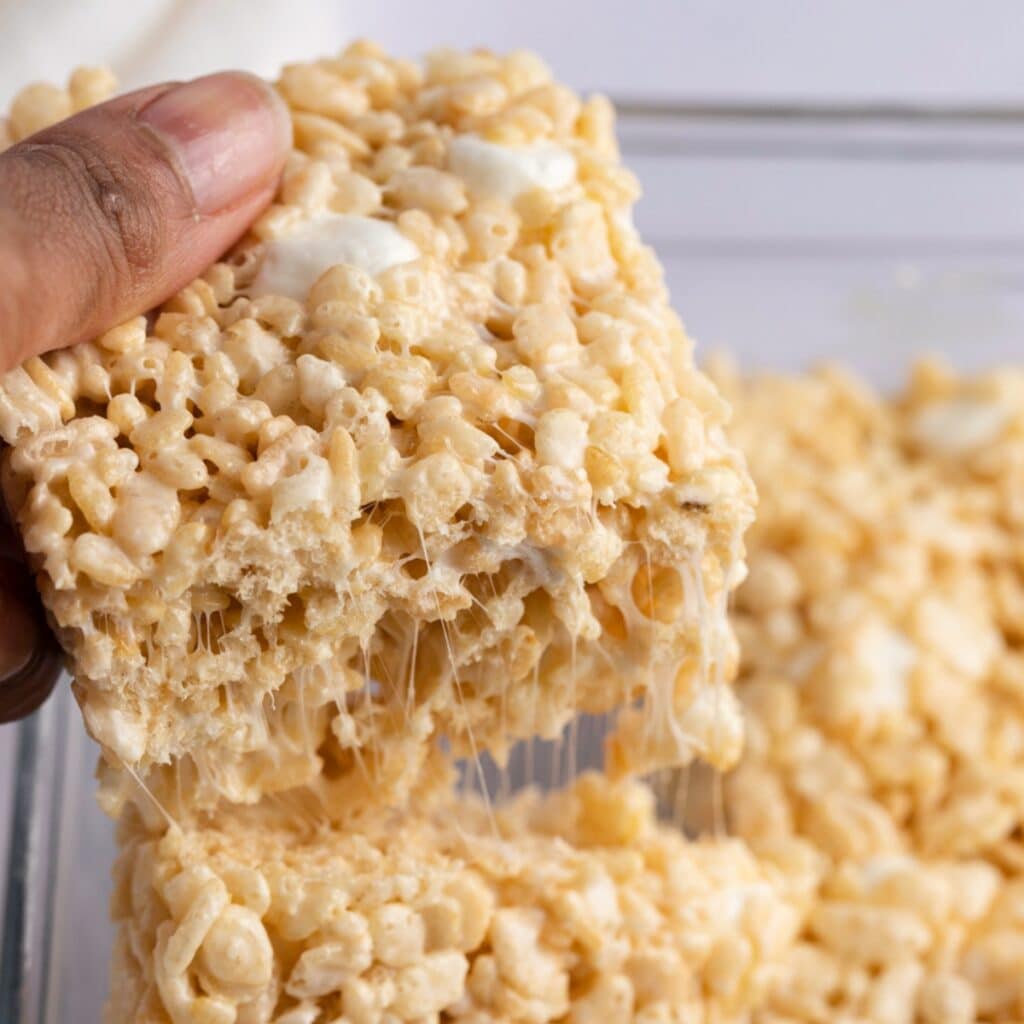 Hand holding a slice of rice krispie treat from a baking dish. 