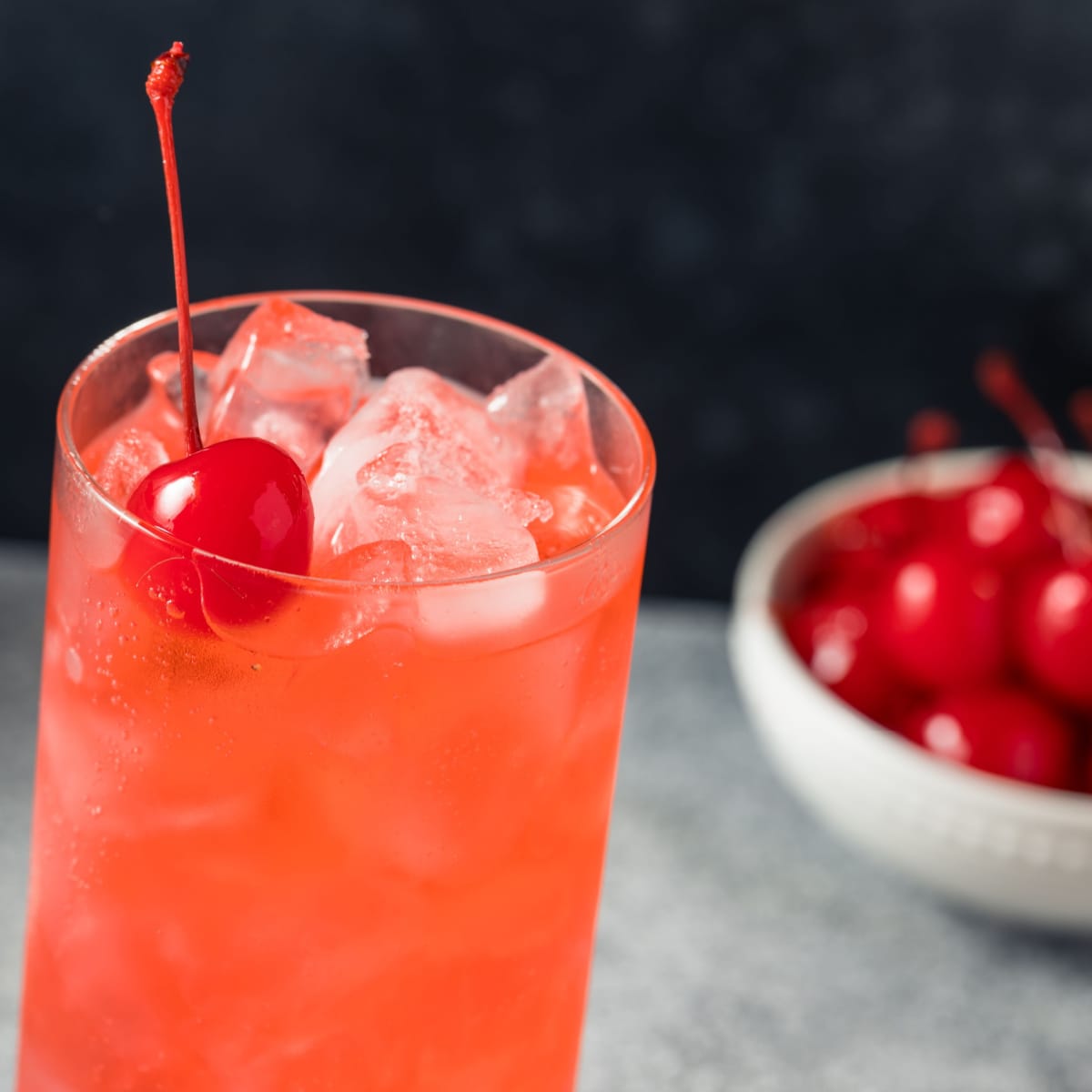 Shirley Temple cocktail served on a tall glass filled with ice and a bowl of fresh cherries in the background. 