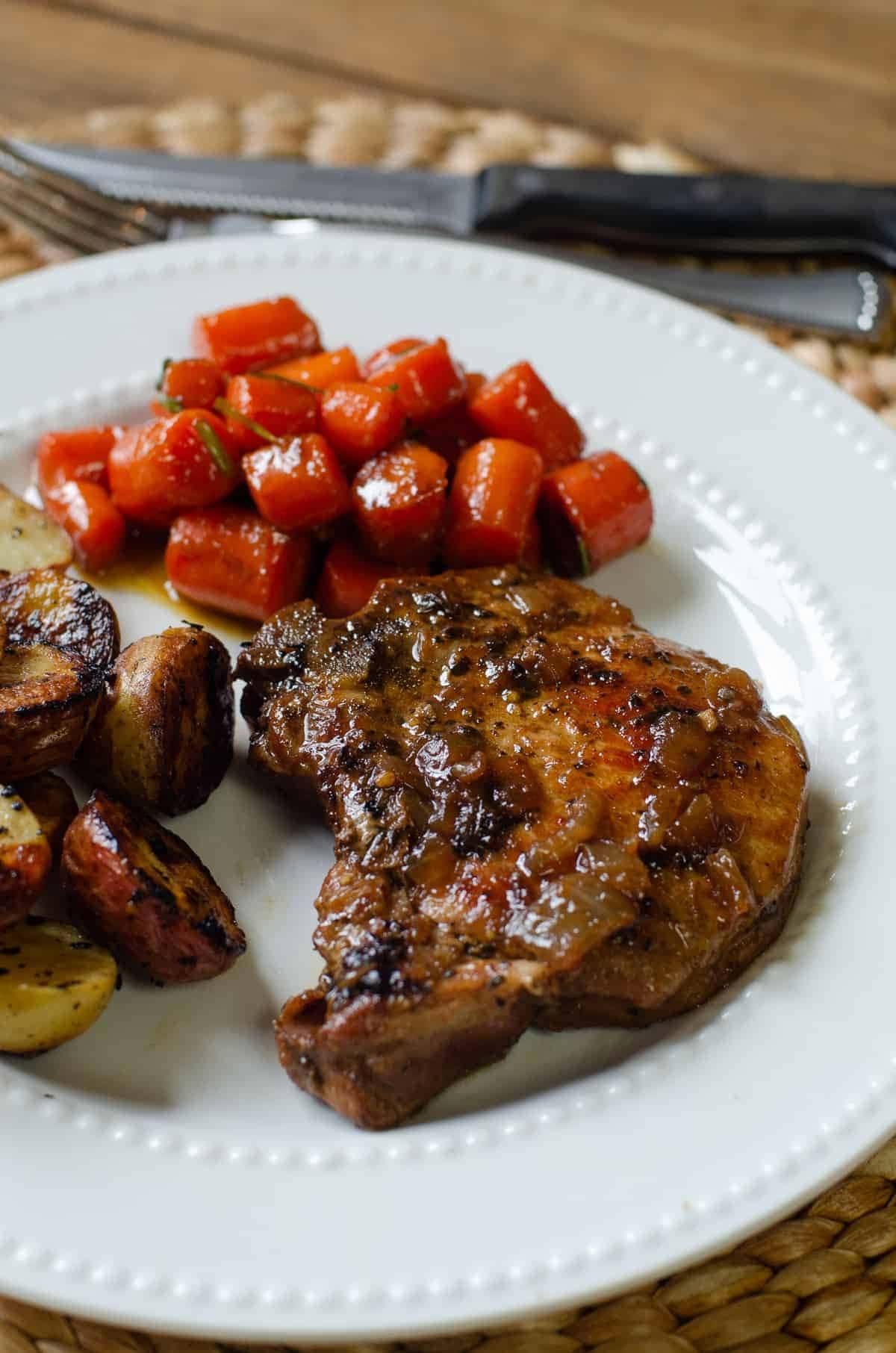 Skillet braised pork chop served on a white plate with carrots and potatoes on the sides. 